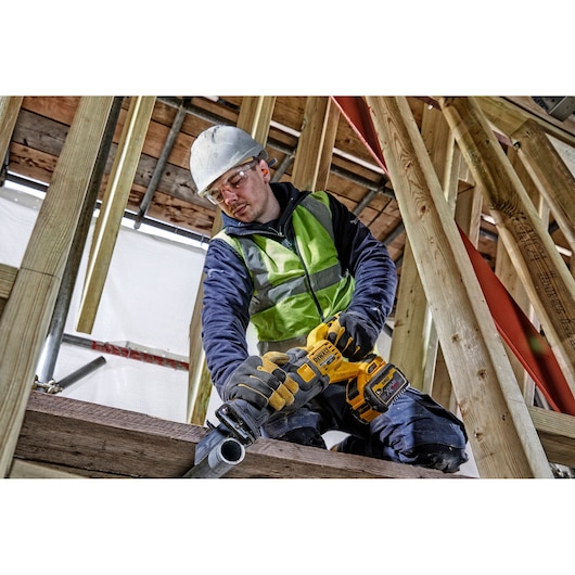 Worker uses a DEWALT cordless reciprocating saw to cut metal pipe at a construction site with wooden framing.