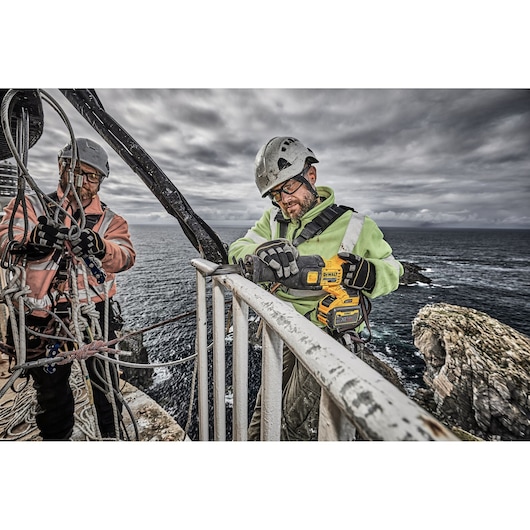 Two workers with safety gear using a DEWALT power tool on a railing, by the sea on a cloudy day.