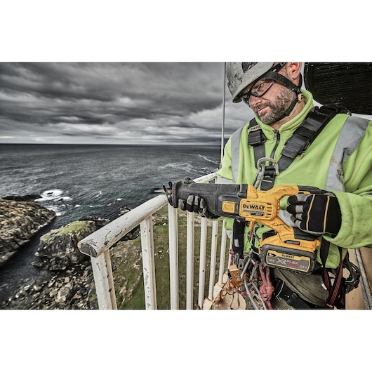Worker holding a DEWALT DCS389X2 reciprocating saw outdoors near a coastline, wearing safety gear and gloves.