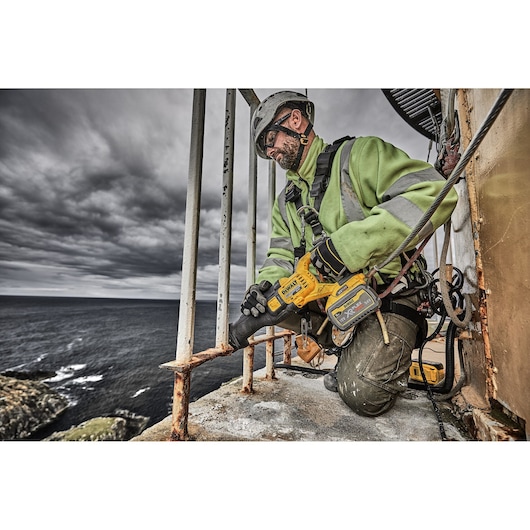 Worker uses DEWALT cordless saw to cut rusty metal railing outdoors, near ocean, wearing safety gear.