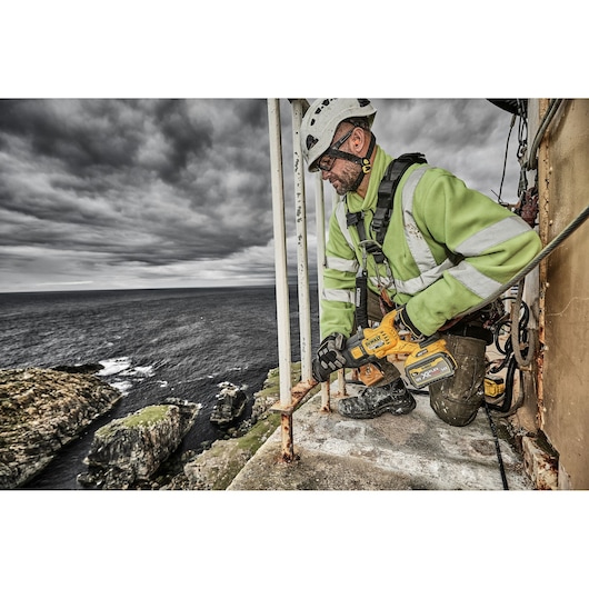 Worker using DEWALT saw on metal railing by a cliffside, wearing safety gear and high-vis jacket.