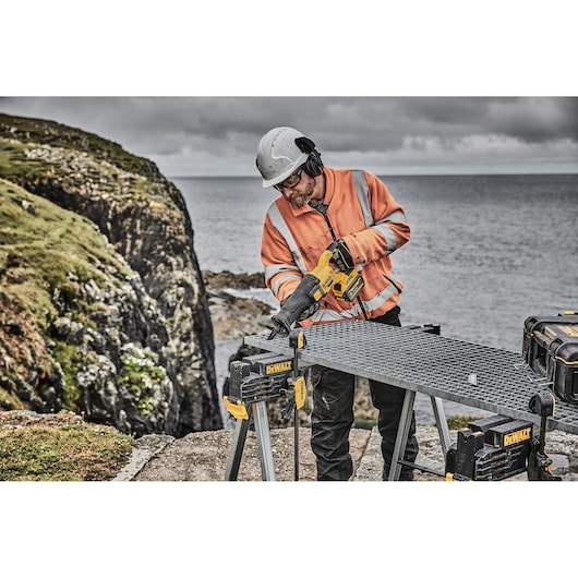 Worker using DEWALT DCS389X2 saw on a metal workbench outdoors by the sea, wearing safety gear.