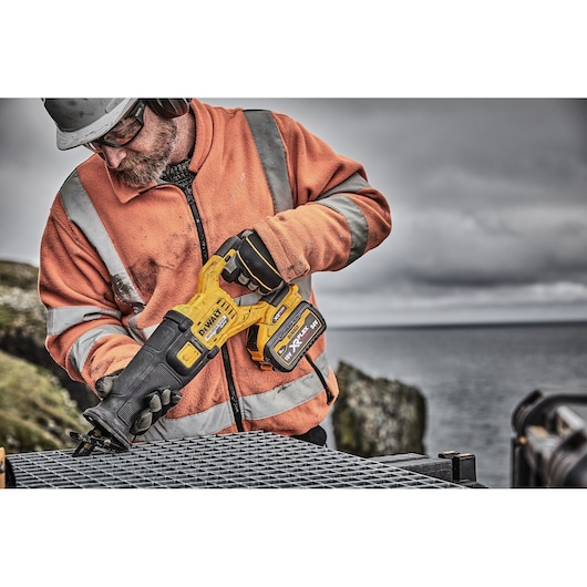 Worker holding a DEWALT reciprocating saw outdoors, cutting metal grid, wearing orange safety jacket.
