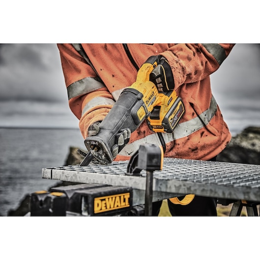 Person uses a DEWALT cordless reciprocating saw to cut metal outdoors, wearing a high-visibility jacket.