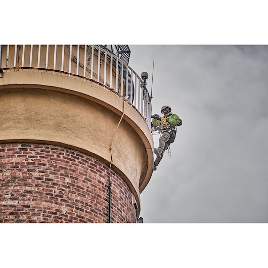 Worker using safety harness drills on side of a brick building near railing, cloudy sky in background.