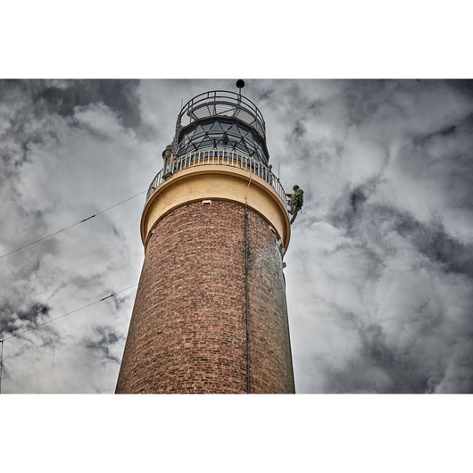 Worker climbs near the top of a tall brick lighthouse against a cloudy sky.