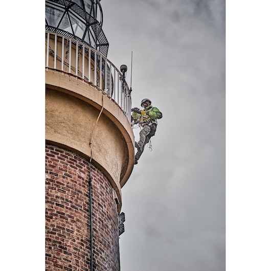 Worker in safety gear uses tools while suspended beside a brick lighthouse against a cloudy sky.