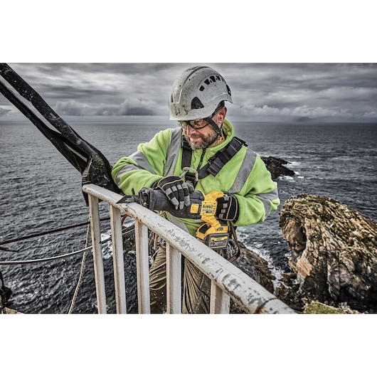 Worker in safety gear uses a DEWALT DCS389X2 saw near a railing by the sea, with rocks and cloudy sky in background.