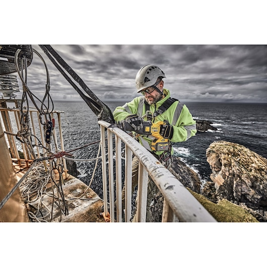 Worker uses a DEWALT DCS389X2 saw outdoors, wearing safety gear on a coastal cliff.