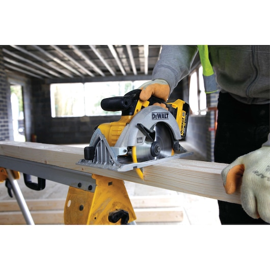A person using a DEWALT 18V cordless circular saw to cut a wooden plank on a workbench inside a construction area.