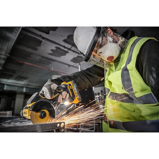 A person wearing safety gear, including a helmet, face shield, mask, gloves, and a high-visibility vest, is using a DEWALT cordless metal cutting tool, generating sparks while working on a metal surface in an industrial environment.
