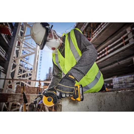 A worker wearing a safety helmet, face shield, mask, high-visibility vest, and DEWALT-branded gloves uses a DEWALT DCS438E2T cordless tool to cut through concrete at a construction site.