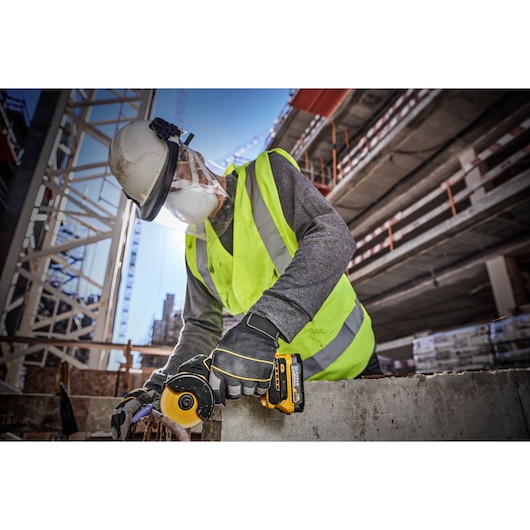 A construction worker wearing a safety helmet, high-visibility vest, gloves, and face shield is using a DEWALT DCS438N cordless tool to cut or grind concrete at a building site.