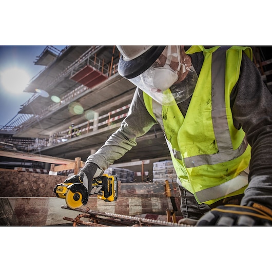 A construction worker wearing a high-visibility vest, protective gloves, a face mask, and a helmet uses a DEWALT DCS438N cordless power tool to cut metal rebar on a construction site with a partially built structure in the background.