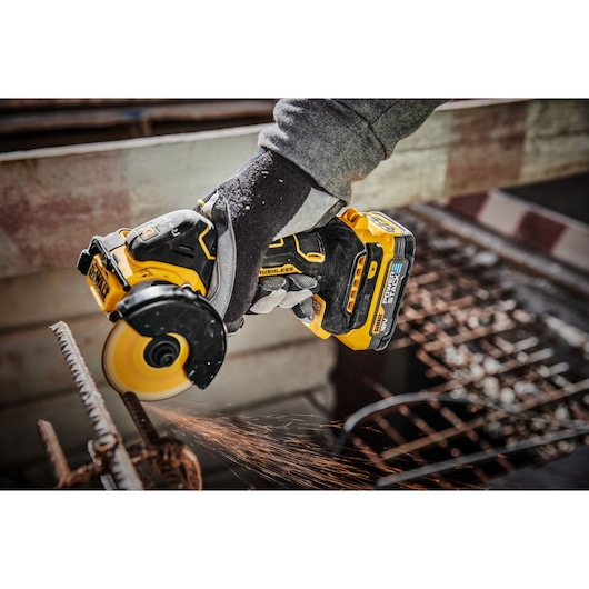 A close-up of a person wearing gloves using a DEWALT DCS438N cordless brushless power tool to cut through a metal rod, with visible sparks flying from the cutting process.
