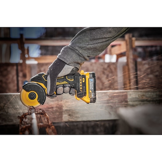 A person wearing work gloves is using a DEWALT DCS438N cordless brushless angle grinder to cut a metal rod, with sparks flying in an industrial or construction setting.