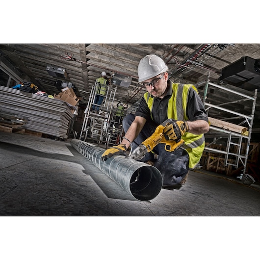 Worker using a DEWALT cordless shear to cut a metal duct pipe on a construction site.