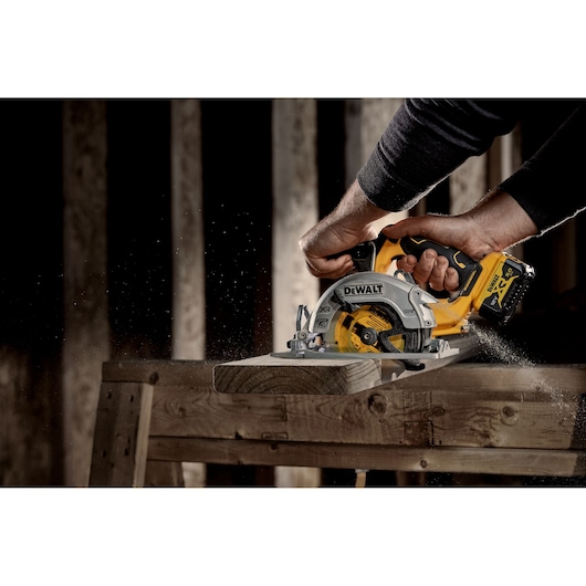 Person using a DEWALT cordless circular saw to cut wood on a workbench with sawdust flying.