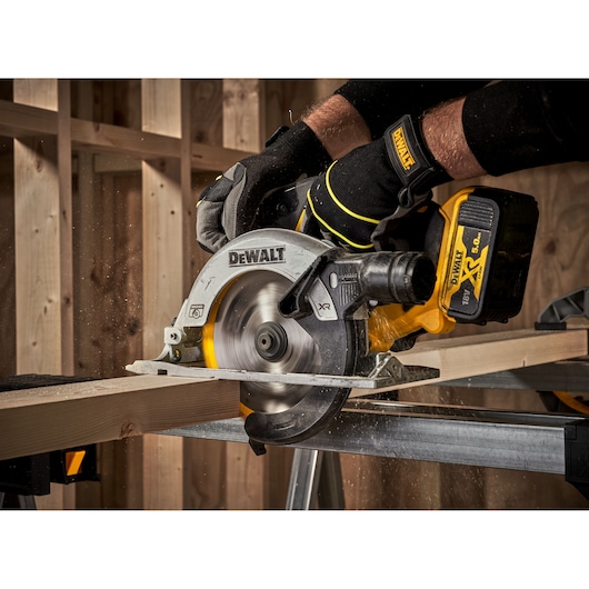 DEWALT cordless circular saw cutting wood, held by gloved hands in a workshop.