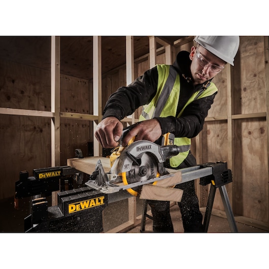 Person using a DEWALT circular saw to cut wood on a worksite, wearing safety gear and high-visibility vest.