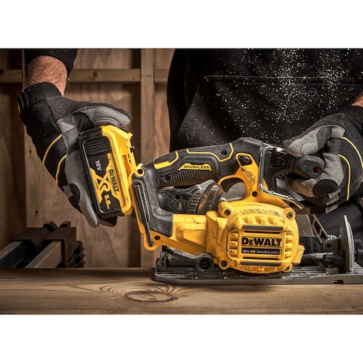 Gloved hands insert battery into DEWALT 18V brushless circular saw on wooden surface, close up view.