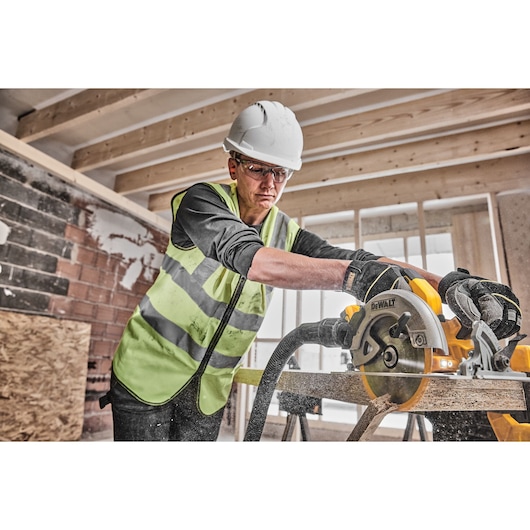 Worker uses DEWALT circular saw to cut wood at construction site, wearing safety gear and gloves.