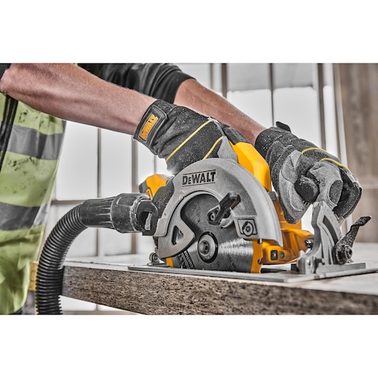 Gloved hands using a DEWALT cordless circular saw to cut wood, with a vacuum attachment.