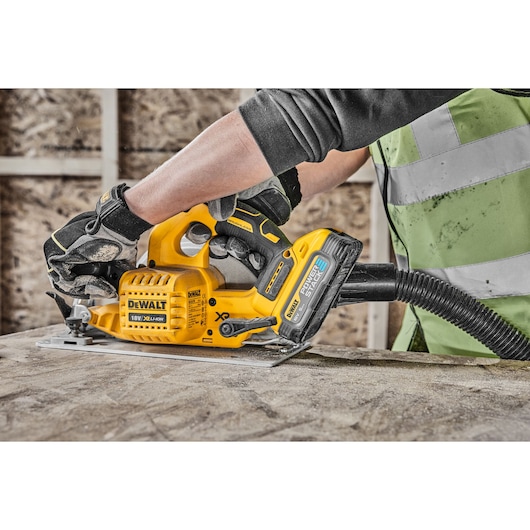 Person uses a DEWALT cordless circular saw with dust extraction on a wooden surface at a construction site.
