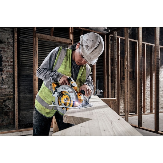 A person wearing a safety vest and hard hat using a DEWALT DCS573H2T cordless circular saw to cut a large wooden plank in an unfinished construction site.