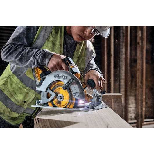 A person wearing safety gear is using a DEWALT cordless circular saw to cut a wooden beam in a workshop or construction site.