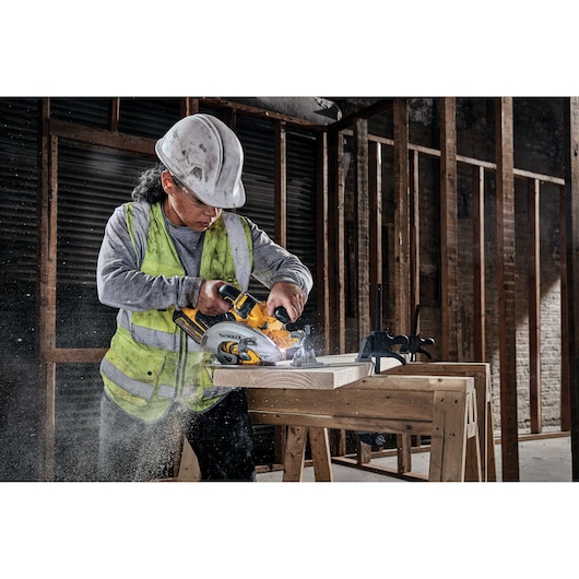 Construction worker using a DEWALT circular saw to cut wood on a workbench in an unfinished building.
