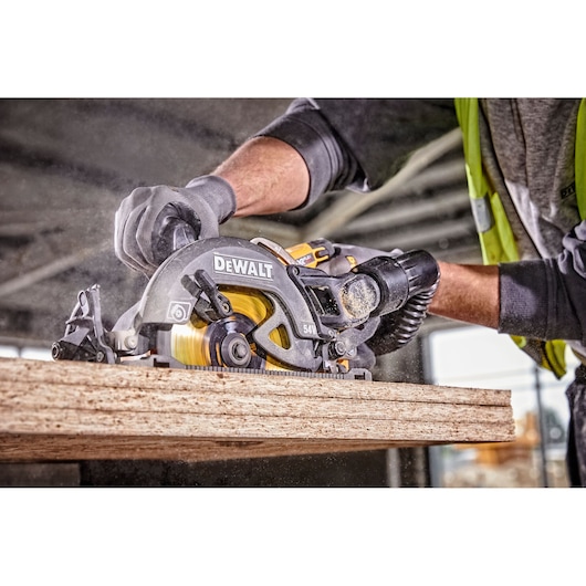Worker using a DEWALT circular saw to cut wood, wearing gloves and safety gear, indoors.