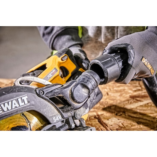Hands connect a dust extractor to a DEWALT circular saw, shown on a wooden surface during work.