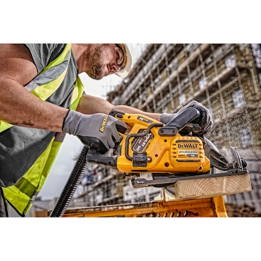 Worker using a DEWALT cordless saw to cut wood on a construction site, wearing gloves and a safety vest.