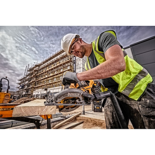 Worker using a DEWALT DCS577T2 circular saw to cut wood at a construction site, wearing safety gear.