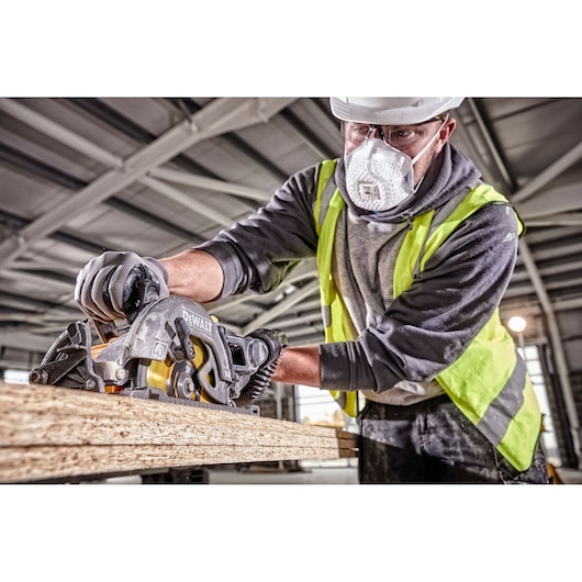 Worker using a DEWALT DCS577T2 power saw to cut wood indoors, wearing gloves and a high-visibility vest.
