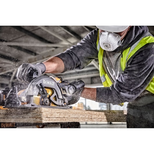 Worker wearing safety gear uses a DEWALT circular saw to cut wood, creating sawdust in a construction setting.