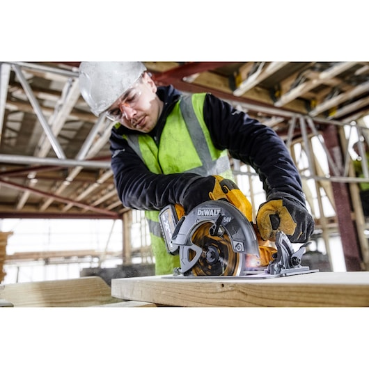DEWALT DCS578NT circular saw cutting wood on a construction site, worker in safety gear.