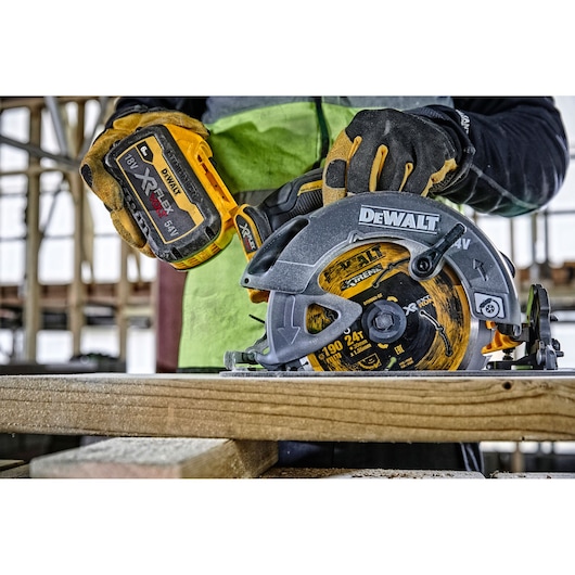 Worker uses DEWALT cordless circular saw to cut wood in a construction setting, wearing gloves for safety.