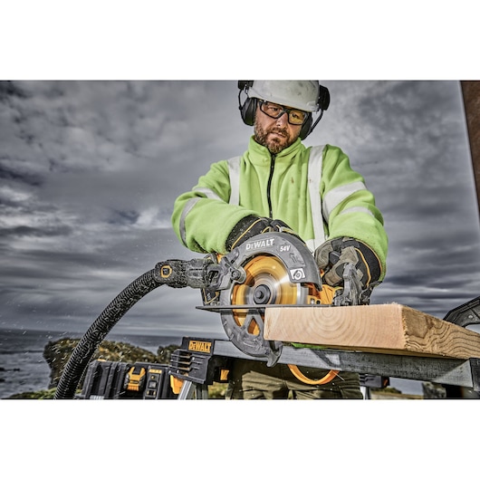Person using DEWALT DCS578X2 circular saw to cut wood outdoors, wearing safety gear and high-visibility jacket.
