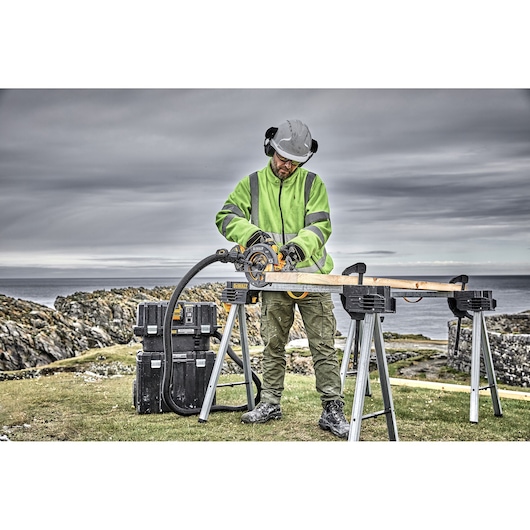 Person using DEWALT circular saw to cut wood outdoors on sawhorses, with tool boxes and rocky landscape in background.