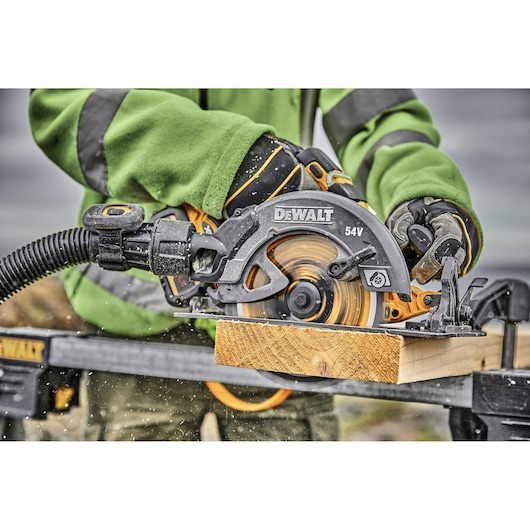 DEWALT 54V circular saw in use, cutting wood with dust extraction attachment, worn by a person in green clothing.
