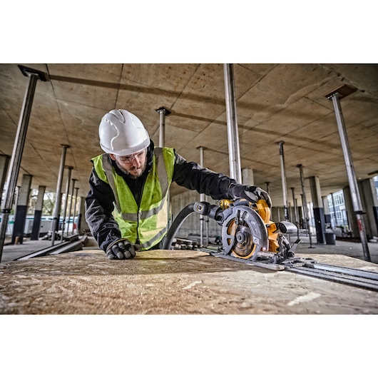 Worker using DEWALT DCS579T2 circular saw on wood panel at a construction site, wearing safety gear.