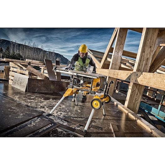 Worker uses DEWALT DCS7485N table saw on construction site with wooden beams and safety gear.