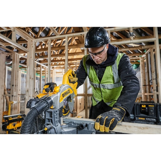 Worker uses DEWALT DCS777T2 saw to cut wood at a construction site, wearing safety gear and gloves.