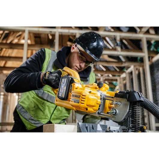 Worker using a DEWALT cordless saw DCS777T2 in a wooden construction area, wearing safety gear and vest.