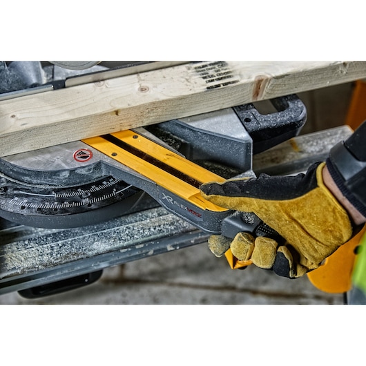 Gloved hand adjusts a DEWALT miter saw cutting a wooden plank. Sawdust covers the tool and work area.