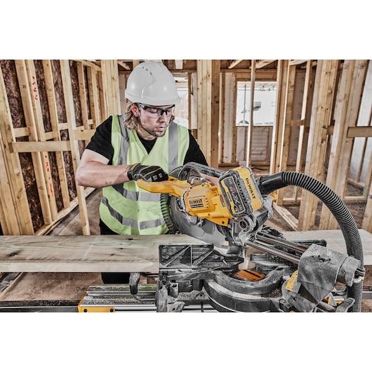 Worker using a DEWALT DCS781N saw to cut wooden planks at a construction site while wearing safety gear.