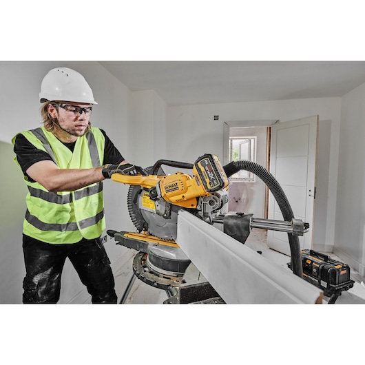 Worker uses DEWALT DCS781N cordless miter saw to cut wood indoors, wearing safety gear.