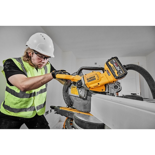 Person using a DEWALT DCS781N battery-powered miter saw on a construction site, wearing safety equipment.